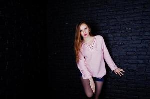 studio shot van brunette meisje op roze trui met jeans shorts tegen zwarte bakstenen muur. foto