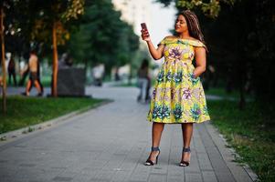 schattig klein Afrikaans Amerikaans meisje met dreadlocks, draag bij gekleurde gele jurk, poseerde bij zonsondergang, kijkend naar telefoon. foto