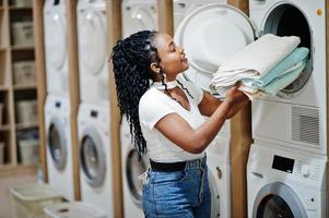 vrolijke Afro-Amerikaanse vrouw met handdoeken in handen in de buurt van wasmachine in de zelfbedieningswasserette. foto