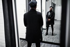 stijlvolle casual Afro-Amerikaanse man op zwarte baret en overjas met heuptas bij paskamer kledingwinkel, kijkend op spiegel. foto