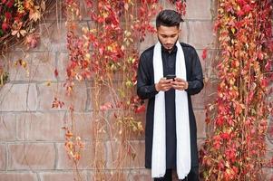 indiase stijlvolle man in zwarte traditionele kleding met witte sjaal poseerde buiten tegen de muur van rode bladeren en keek naar mobiele telefoon. foto