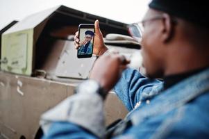 Afro-Amerikaanse man in spijkerjasje, baret en bril, rokende sigaar en poseerde tegen btr militair gepantserd voertuig, selfie makend op de telefoon. focus op het scherm van de mobiele telefoon. foto