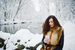 elegantie krullend meisje in bontjas met mobiele telefoon in besneeuwde forest park in de winter. foto