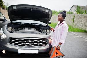 jonge trieste afrikaanse vrouw staat met nooddriehoek tegen auto met gepofte kap. transport, voertuigen problemen en storingen concept. foto