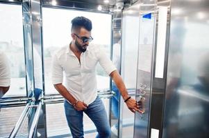 stijlvolle lange Arabische man model in wit overhemd, jeans en zonnebril poseerde bij lift binnen. foto