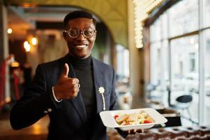 modieuze Afro-Amerikaanse man in pak en bril staan in café met bord salade in de hand en duim opdagen. foto