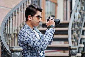 stijlvolle indiase jongeman fotograaf bij zonnebril draagt casual buiten tegen ijzeren trappen met dslr fotocamera bij de hand. foto