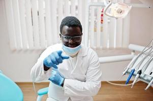 Afro-Amerikaanse mannelijke arts in masker en bril met gekruiste armen zittend op tandartsstoel in tandheelkundige kliniek en vinger naar camera tonen. foto