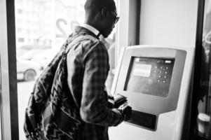 Afro-Amerikaanse man in geruit overhemd met rugzak portemonnee in de hand houden tegen ATM. zwarte man reiziger. foto