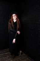 studio shoot van meisje in het zwart met dreadlocks, op bril en hoed op baksteen achtergrond. foto