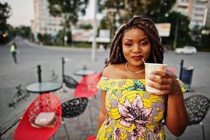 schattig klein Afrikaans Amerikaans meisje met dreadlocks, draag bij gekleurde gele jurk, poseerde op terras met kopje koffie. foto