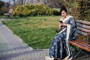 elegante brunette Zuid-Aziatische Indiase meisje in saree zittend op de bank met mobiele telefoon bij de hand. foto