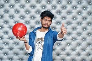 stijlvolle aziatische man in jeans shirt staande met bowlingbal bij de hand tegen zilveren muur achtergrond en duim opdagen. foto