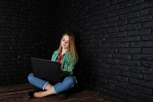 stijlvol blond meisje in jas en spijkerbroek met laptop tegen zwarte bakstenen muur in de studio. foto