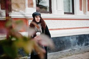 krullend mexicaans meisje in leren pet en plastic kopje koffie bij de hand wandelen in de straten van de stad. foto