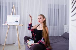 mooie vrouw kunstenaar schilder met borstels en olie canvas poseren bed op kamer. foto