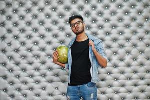 stijlvolle Aziatische man in jeans jasje en bril met bowlingbal bij de hand tegen zilveren muur achtergrond. foto