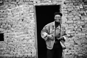 Afro-Amerikaanse man in jeans jasje, baret en bril tegen bakstenen muur op verlaten dak. foto