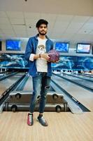 stijlvolle baard Aziatische man in jeans shirt staande op bowlingbaan met bal bij de hand. foto