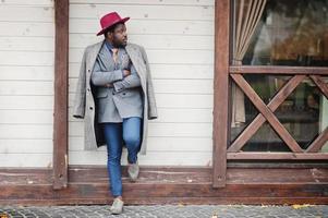 stijlvolle Afro-Amerikaanse man model in grijze jas, jas stropdas en rode hoed gesteld tegen houten café. foto