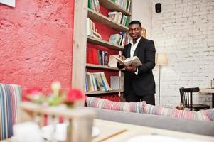 sterke krachtige afro-amerikaanse man in zwart pak lees boek tegen planken in gratis boeken anti-café. foto