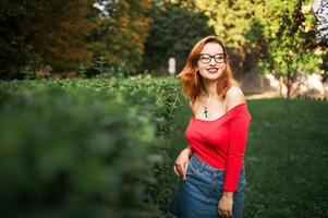 aantrekkelijke roodharige vrouw in brillen, dragen op rode blouse en jeans rok poseren in groen park. foto