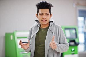 jonge stijlvolle aziatische man met creditcard in de hand toont duim tegen rij groene geldautomaat. foto