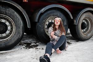 brunette stijlvolle casual meisje in pet en zonnebril zittend tegen vrachtwagen wielen. foto