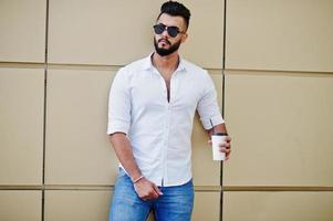 stijlvolle lange Arabische man model in wit overhemd, jeans en zonnebril gesteld op straat van de stad. baard aantrekkelijke Arabische man met kopje koffie tegen gouden muur. foto