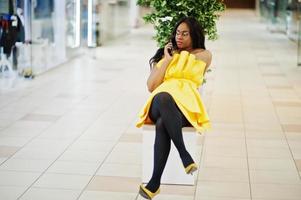 stijlvolle Afro-Amerikaanse vrouw in gele jurk poseerde in het winkelcentrum en zat op een bankje binnen, aan de telefoon. foto