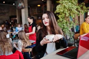 brunette meisje zittend op café met kopje cappuccino, werken met rode laptop. foto