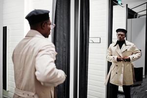 stijlvolle casual Afro-Amerikaanse man bij zwarte baret en witte trenchcoat bij paskamerkledingwinkel, kijkend naar spiegel. foto