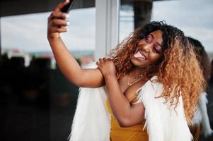 glamour Afro-Amerikaanse vrouw in gele jurk en witte wollen cape selfie maken op de telefoon. foto