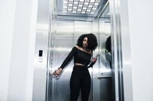 modieuze jonge mooie Afro-Amerikaanse zakenvrouw met afro kapsel dragen in elegant zwart, staan bij de lift met telefoon en laptop bij de hand. foto