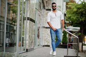 stijlvolle lange Arabische man model in wit overhemd, jeans en zonnebril gesteld op straat van de stad. baard aantrekkelijke Arabische man. foto