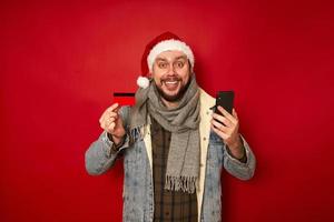 opgewonden man in kerstmuts houdt bankcreditcard vast en koopt telefoon via mobiele applicatie in online winkel geschenken voor nieuwjaar geïsoleerd rode studio achtergrond concept - mensen, technologie, winkelen foto