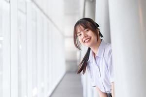 mooie middelbare school Aziatische student meisje in het schooluniform staat en glimlacht gelukkig terwijl ze vol vertrouwen naar de camera kijkt met het schoolgebouw als achtergrond. foto