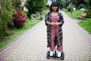 mooie afrikaanse amerikaanse vrouw die segway of hoverboard gebruikt. zwarte meid op zelfbalancerende elektrische scooter met twee wielen met mobiele telefoon bij de hand. foto