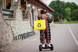 mooie afrikaanse amerikaanse vrouw die segway of hoverboard en mobiele telefoon gebruikt. zwart meisje met gele stoffen eco-tassen recycling symbool. foto