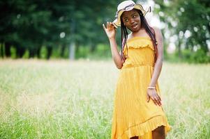 portret van prachtige Afro-Amerikaanse vrouw 20s in slijtage in gele jurk en zomerhoed poseren op groen gras in park. foto