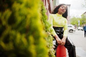 portret van jonge mooie Afro-Amerikaanse fashion model vrouw, draag in groene top en zwarte glanzende natte look broek, zittend op rood vat. foto