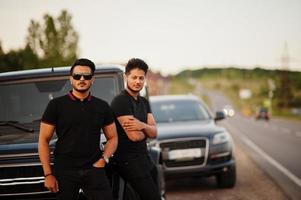 twee aziatische broers die een man dragen op alle zwarte poses in de buurt van suv-auto's. foto