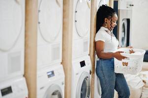 vrolijke Afro-Amerikaanse vrouw met witte mand in de buurt van wasmachine in de zelfbedieningswasserette. foto