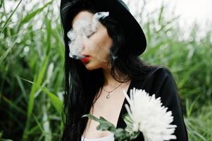 sensueel rokersmeisje helemaal in zwarte, rode lippen en hoed. goth dramatische vrouw houdt witte chrysantenbloem vast en rookt op gewoon riet. foto
