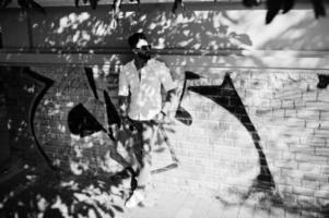 stijlvolle lange Arabische man model in wit overhemd, jeans en zonnebril gesteld op straat van de stad. baard aantrekkelijke arabische man tegen graffiti muur. foto