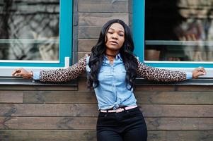 hipster afrikaans amerikaans meisje met een jeansoverhemd met luipaardmouwen die zich voordeed op straat tegen een houten huis met ramen. foto
