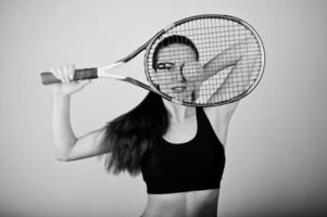 zwart-wit portret van een mooie jonge vrouw in sportkleding die een tennisracket vasthoudt terwijl hij tegen een witte achtergrond staat. foto