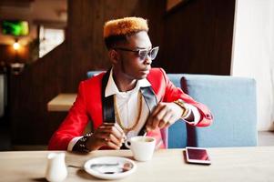 mode Afro-Amerikaanse man model in rood pak, met hoogtepunten haar en zonnebril zittend in café met koffie. foto