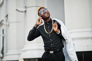 chique knappe Afro-Amerikaanse man in wit pak en zwart shirt met mobiele telefoon. foto