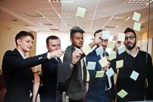 business mans wijzend op glas met kleurrijke papieren notities. diverse groep mannelijke werknemers in formele kleding met stickers. foto
